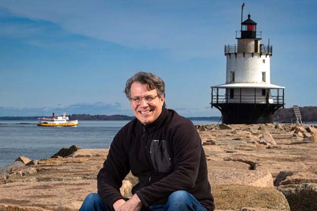 Maine artist, Dominic White sitting on Spring Point Light jetty with lighthouse and ferry behind him.